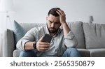 Psychological difficulties, relationship problems, depression and crisis. Unhappy disappointed sad bearded young man sit on floor hold his head with hands and look at phone, in living room interior