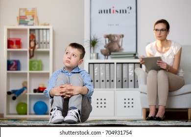 Psychologist In Glasses Is Looking At Kid Sick Of Autism Sitting On Carpet In Classroom. Autistic Child Therapy Concept