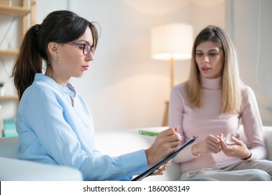 The Psychologist Listens Attentively To Young Woman And Writes In Clipboard While Sitting On Couch.