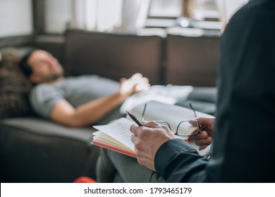 Psychotherapist and patient at an appointment in the office. Doctor's hands with notepad close-up. The doctor records the client's history. A male patient lies on a sofa in a large, bright office