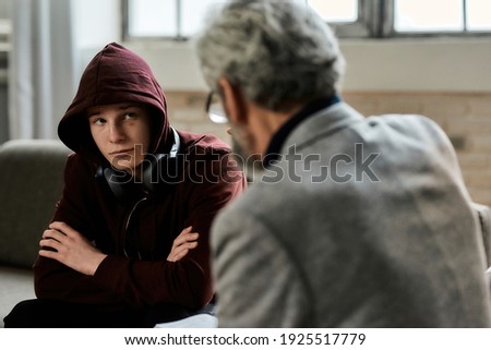Psychotherapist talking to troubled teenage boy in hood sitting with crossed hands on couch during appointment in office, selective focus. Psychotherapy concept