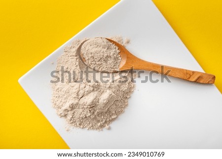Psyllium husk flour powder on white plate and wooden spoon indoors at home, yellow background. Health benefits of Psyllium flour concept.