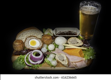 Ploughman'?s Pub Lunch Including Scotch Egg; Pork Pies, Ham; Pickled Onions And Cheese Served With A Pint Of Lager Beer. Isolated Against A Black Background.