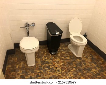 Public Bathroom With Two White Toilets And No Partition. One Kid Is Open And One Is Closed. With A Black Garbage Can In Between. Brown Tiled Floor. White Tiled Walls