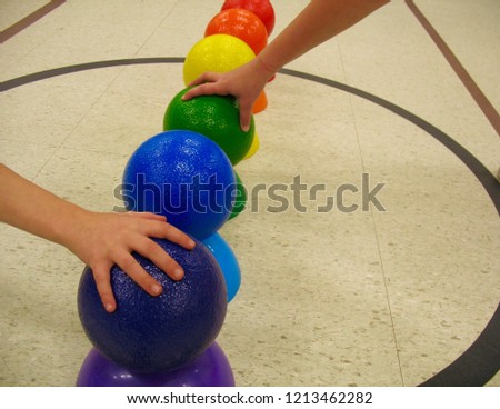 In a public, crowded, fun gymnasium, under fluorescent lights, are colorful dodge balls, perched upon a matching cone, ready for a child to grab and throw them into action from the mid-court circle