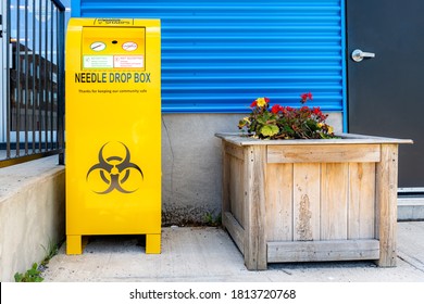 A public drop box for used needles, on a sidewalk by a building. The box is bright yellow, and bears the standard bio-hazard symbol.