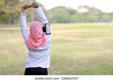 Public Exercise Lifestyle Concept, Fitness Woman Listening Music From Wireless Earphones And Mobile Phone. Rear View Athletic Young Asian Girl In Muslim Sportswear Standing, Stretching After Workout 