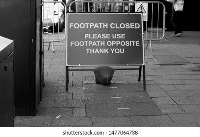 A Public Information Sign Stating: Footpath Closed Please Use Footpath Opposite Thank You, With The Display Frame Held Down By A Single Sandbag On A Paving Slab Pavement. 