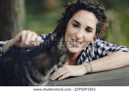 In a public park, a woman in a black-and-white plaid shirt is petting and cuddling a big cat she found. He enjoys the pleasure because cats know how to do that.