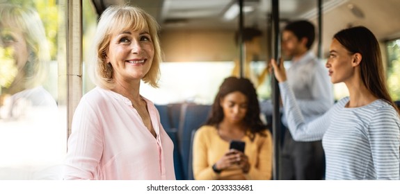 Public Transportation Concept. Portrait Of Smiling Senior Lady Standing In Bus, Leaning Looking Out Of Window, Going Home, Banner, Panorama. Group Of Diverse People Passengers In Blurred Background