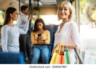 Public Transportation. Group of diverse people taking bus, selective focus on smiling mature woman holding shopping bags, standing and looking out of window, leaning on handle going home