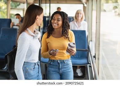 Public Transportaton. Two Multiethnic Smiling Young Women Standing At Autobus And Talking, Black Lady Holding Takeaway Coffee Cup And Cell Phone, Students Enjoying Ride Travel Together Inside Vehicle