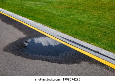 Puddle On Asphalt Road With Stormwater On The Side Of The Road With A Ditch For Water And A Glade Of Green Lawn Along The Road.