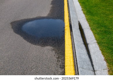 Puddle On Asphalt Road With Stormwater On The Road Side With A Ditch For Water After Rain And A Glade Of Green Lawn Along The Highway.