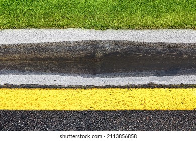 Puddle On Drain Canal Near Asphalt Road With Stormwater On The Road Side With Ditch For Water After Rain And Glade Of Green Lawn Along Highway With Yellow Marking.