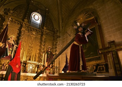 Puerto De Santa María, Spain - Feb. 25, 2022: Ornately Dressed Sculpture Of Jesús Nazareno Carrying The Cross, Golden Baroque Altarpiece And Red Black Brotherhood Of The Nazarene Habit To The Side