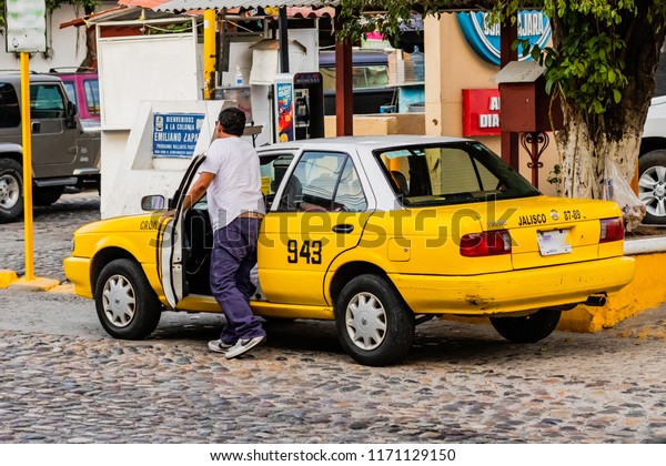 Puerto Vallarta Mexico 02/22/2017 View from across\
street of driver entering a yellow taxi in zona romantica on sunny\
day.