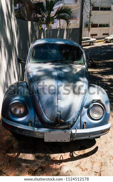 PUERTO VALLARTA, MEXICO - MARCH 10, 2018: An\
oldclassic Volkswagen Beetle, in rough condition, with a yellow\
smiley face hanging in the front window shield, parked in the shade\
off the main road.