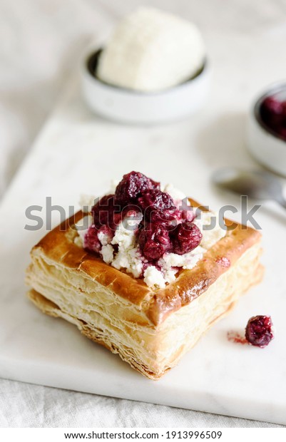 Puff pastry squares with ricotta and cherry sauce\
on white marble tray. Baked small square cakes topped with cheese\
and berry sauce