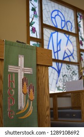 A Pulpit With A Green Banner Depicting A Cross, Wheat And The Word Hope. Blurred In The Background Is A Stained Glass Window Featuring Grapes, Vines And The Outline Of A Shepard.