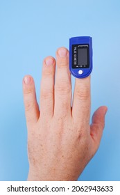 Pulse Oximeter On The Fingertip Of An Elderly Man Isolated On A Blue Medical Background.Measurement, Monitoring Of Pulse Rate, Blood Oxygen Saturation, Lung Testing For Viral Infection.Vertical Photo