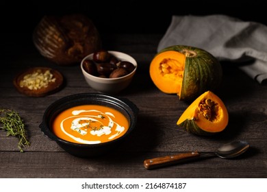 Pumpkin and chestnut soup decorated with cream and thyme aside open pumpkin, chestnuts and bread loaf on a vintage dark wood background. Chiaroscuro light. Autumn concept.