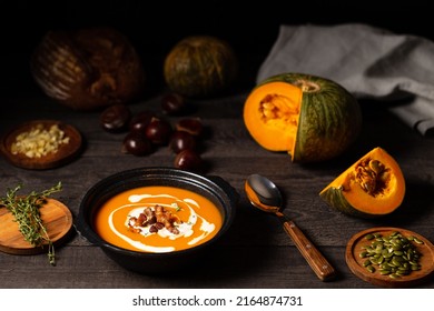 Pumpkin and chestnut soup decorated with cream pork belly and thyme aside open pumpkin, chestnuts and bread loaf on a vintage dark wood background. Chiaroscuro light. Autumn concept.