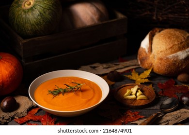 Pumpkin and chestnut soup decorated with rosemary twig aside pumpkins, chestnuts and bread on a vintage dark wood background spread with maple leaves. Chiaroscuro light. Autumn concept.