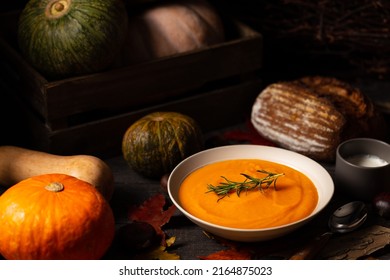 Pumpkin and chestnut soup decorated with rosemary twig aside pumpkins, chestnuts and bread loaf on a vintage dark wood background spread with maple leaves. Chiaroscuro light. Autumn concept.