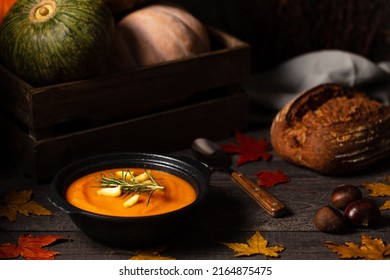 Pumpkin and chestnut soup decorated with rosemary twig aside entire pumpkins in a crate, chestnuts and bread loaf on a vintage dark wood background spread with apple leaves. Chiaroscuro light. Autumn.