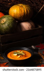 Pumpkin and chestnut soup decorated with rosemary twig and chestnuts aside entire pumpkins in a crate, on a vintage dark wood background spread with apple leaves. Chiaroscuro light. Autumn concept.