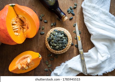 Pumpkin seeds in a bowl, an orange Hokaido pumpkin and oil in a glass bottle, a knife, a cloth napkin on a brown kitchen table. Top view, autumn background.