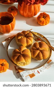 Pumpkin shaped bread buns on a white plate with a golden rim. Surrounded by autumn decorative elements including a pumpkin mug. A pumpkin vase with red flowers. And small velvet pumpkins