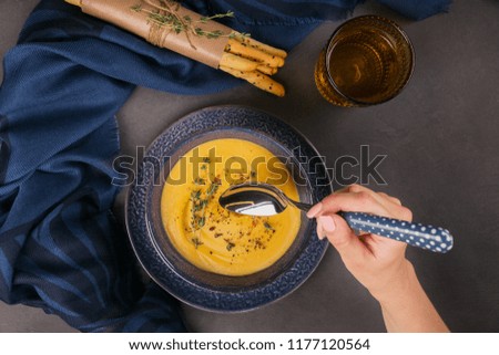 Pumpkin soup in blue bowl topped with fresh herb on dark background. Healthy vegetable food for dietary. Woman's hand holds a spoon