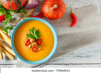 Pumpkin Soup With Chili Pepper, Pumpkin Seeds, Croutons And Cilantro In Blue Bowl On White Wooden Background. Top View, Copy Space