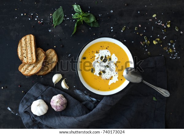 Pumpkin soup with\
cream, seeds, bread and fresh basil in rustic metal plate on grunge\
black background. Top\
view