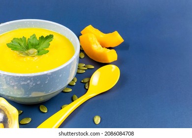 Pumpkin Soup Puree For The Baby In A Beautiful Bowl, Pumpkin Seeds And Pieces, A Yellow Spoon On A Blue Background. Baby Nutrition, The First Complementary Food. Space For Text. Recipe.