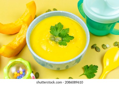 Pumpkin Soup Puree For The Baby In A Blue Bowl, Pumpkin Seeds And Pieces, A Spoon, A Drinking Bowl And A Toy On A Yellow Background. Baby Nutrition, The First Complementary Food.