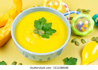 Pumpkin Soup Puree For The Baby, Peeled Pumpkin Seeds And Pieces, A Spoon And A Rattle Toy On A Yellow Background. Close-up. Baby Nutrition, The First Complementary Food.