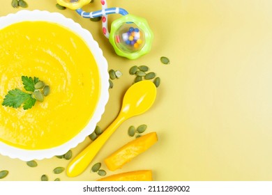 Pumpkin Soup Puree For The Baby, Peeled Pumpkin Seeds And Pieces, A Spoon And A Rattle Toy On A Yellow Background. Baby Nutrition, The First Complementary Food. Place For Text, Recipe.