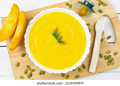 Pumpkin Soup Puree For The Baby, Pumpkin Seeds And Pieces, A Spoon And A Pacifier On A Wooden Board Close-up. Baby Nutrition, The First Complementary Food.