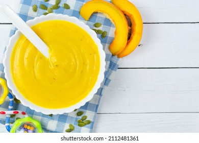 Pumpkin Soup Puree For A Baby With A Spoon, Pumpkin Seeds And Pieces, A Rattle Toy On A Blue Napkin On A White Wooden Background. Place For Text, Recipe. Baby Nutrition, The First Complementary Food.