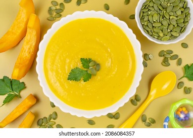 Pumpkin Soup Puree For The Baby In A White Bowl, Pumpkin Seeds And Pieces, A Spoon And A Toy On A Yellow Background. Baby Nutrition, The First Complementary Food. Close-up, Top View.