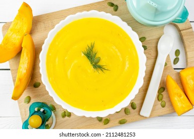 Pumpkin Soup Puree For The Baby In A White Plate, Pumpkin Seeds And Pieces, A Pacifier, A Spoon And A Drinking Bowl On A Wooden Board. Baby Nutrition, The First Complementary Food.