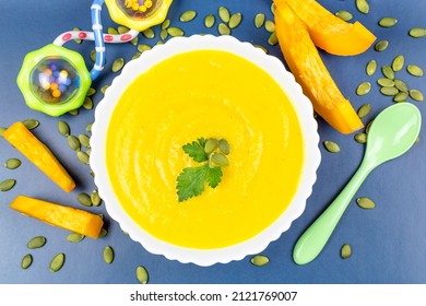 Pumpkin Soup Puree For The Baby In A White Plate, Pumpkin Seeds And Pieces, A Spoon, A Rattle Toy On A Blue Background. Baby Nutrition, The First Complementary Food.