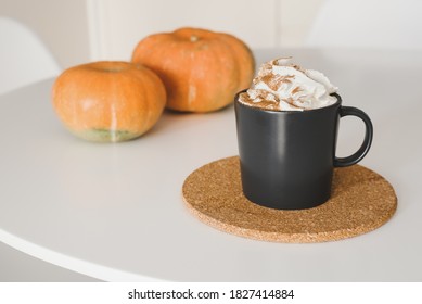 Pumpkin Spice Latte With Whipped Cream And Cinnamon Powder In A Black Coffee Mug Served On Cork Coaster And Two Small Pumpkins At White Table