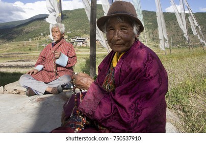 PUNAKHA (BHUTAN) - OCTOBER 2017: Older Couple Praying In The Morning Near A Stupa. Praying Flags Flying In The Background