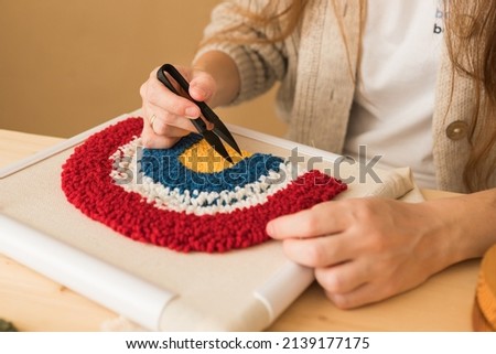 Punch needle embroidery.  woman triming  with scissors the strands threads that sewn through and longer than others on foundation fabric stretch  over the frame.  Handicraft, needlework, hobby, diy