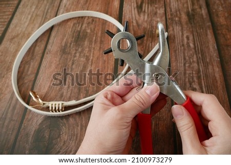 Puncture holes in the strap using a punch and belt on wooden background. Belt Hole Puncher Power Assist Heavy Duty Revolving Eyelet Punch Plier. Closeup