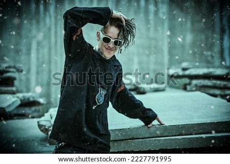 Punk guy. Rock culture. An extraordinary mature man with dreadlocks and tattoos wearing black clothes with ethnic ornaments smiles joyfully standing on the street under falling snowflakes.